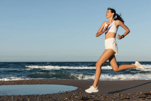 Full length of cheerful young sportive woman jogging near sea in summer — Stock Photo