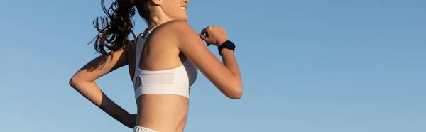 Cropped view of cheerful young and sportive woman jogging against blue sky, banner — Stock Photo