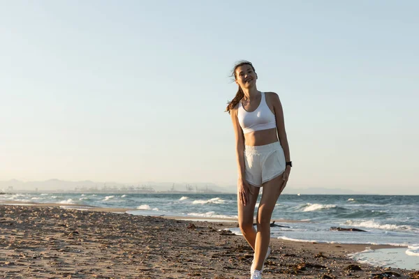 Happy woman in white sports bra and shorts standing near sea on beach — Stock Photo