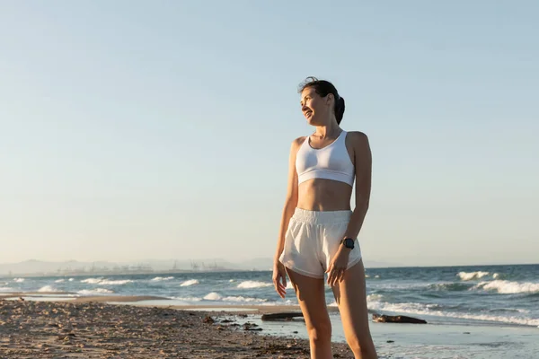 Donna allegra in reggiseno sportivo bianco e pantaloncini in piedi vicino al mare sulla spiaggia — Foto stock
