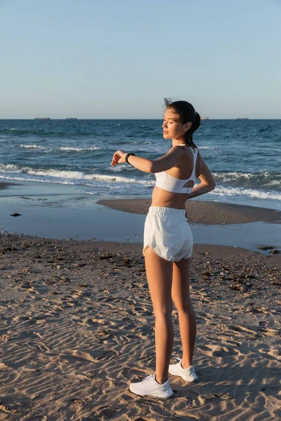 Jovem mulher em sutiã esportivo e shorts olhando para rastreador de fitness perto do mar na praia — Fotografia de Stock