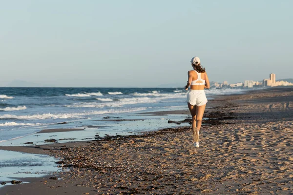 Vista posteriore della giovane donna sportiva in berretto da baseball che fa jogging vicino al mare in estate, banner — Foto stock