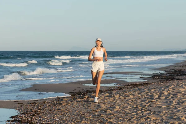 Cheerful young sportive woman in baseball cap jogging near sea in summer — Stock Photo