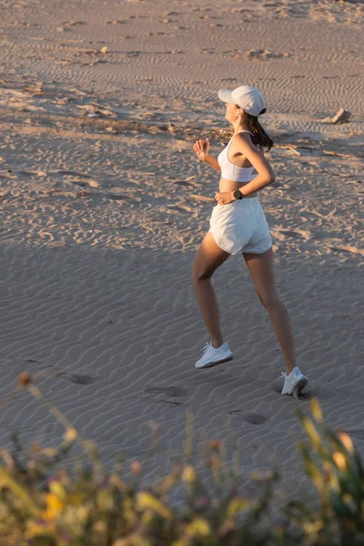 Jeune femme sportive en casquette de baseball et écouteur sans fil fonctionnant sur sable — Photo de stock