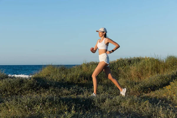 Mulher esportiva em boné de beisebol e fone de ouvido sem fio correndo na grama perto do mar — Fotografia de Stock