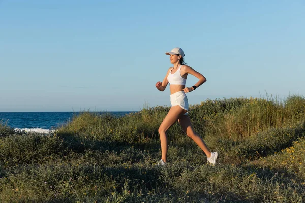Sportive woman in baseball cap and wireless earphone running on grass near sea — Stock Photo