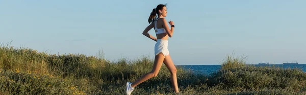 Jovem mulher esportiva em shorts e fone de ouvido sem fio correndo na grama perto do mar, bandeira — Fotografia de Stock