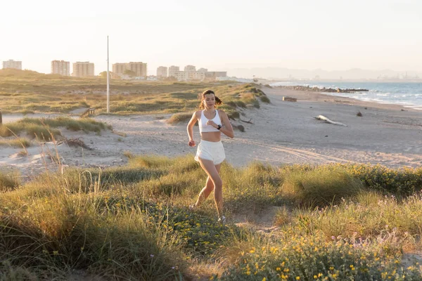 Joven mujer deportiva en pantalones cortos y auriculares inalámbricos trotando en la hierba cerca del mar - foto de stock