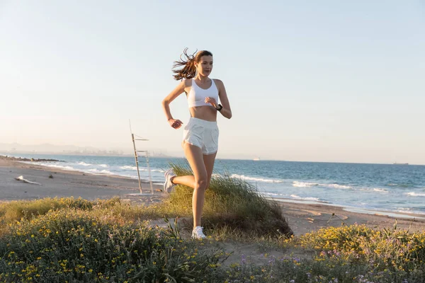 Jovem mulher em shorts e fone de ouvido sem fio jogging na grama perto do mar — Fotografia de Stock