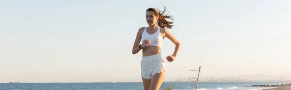 Mujer joven en pantalones cortos y auriculares inalámbricos corriendo cerca del mar, pancarta - foto de stock