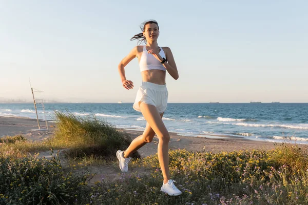 Sorrindo jovem mulher em shorts e fones de ouvido sem fio correndo na grama perto do mar — Fotografia de Stock