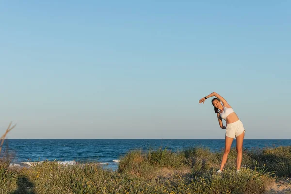 Jovem mulher em shorts esportivos e fones de ouvido sem fio que se estendem perto do mar azul — Fotografia de Stock