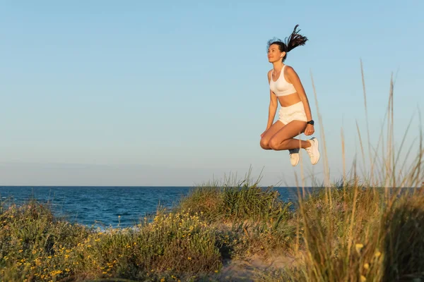 Mujer joven en pantalones cortos deportivos y auriculares inalámbricos saltando cerca del mar azul - foto de stock