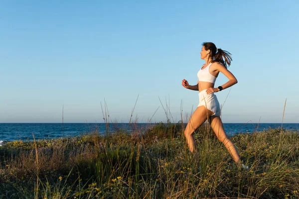 Vue latérale de la jeune femme en short et écouteurs sans fil jogging sur l'herbe près du rivage de la mer — Photo de stock