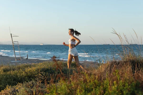 Vista lateral da jovem mulher em sutiã esportivo e fone de ouvido sem fio jogging na grama perto da costa do mar — Fotografia de Stock