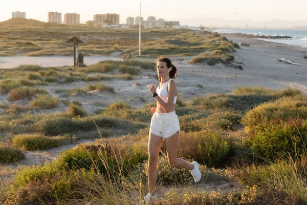 Mujer en forma en pantalones cortos y sujetador deportivo escuchar música y correr cerca de la orilla del mar - foto de stock