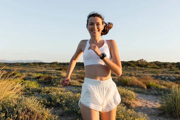 Pleased woman in shorts and sports bra listening music and running outside — Stock Photo