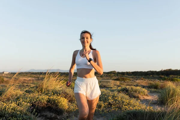 Smiling woman in shorts and sports bra listening music and running outside — Stock Photo