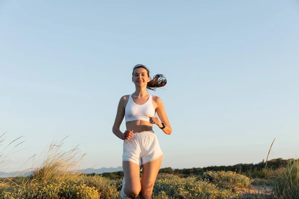 Deportista feliz en pantalones cortos y sujetador deportivo escuchar música y correr fuera - foto de stock