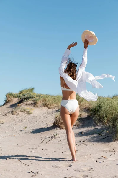 Vue arrière de la femme brune tenant un chapeau de soleil et marchant sur le sable — Photo de stock