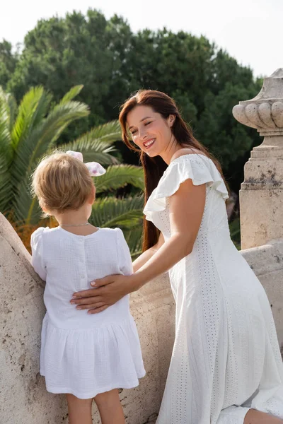 Happy woman in summer dress hugging daughter on balcony of stone building in Valencia - foto de stock