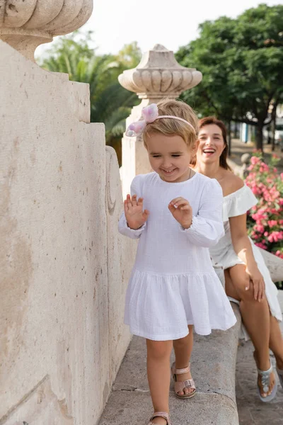 Smiling child walking on stone bench of Puente Del Mar bridge near blurred mom in Valencia — Stock Photo