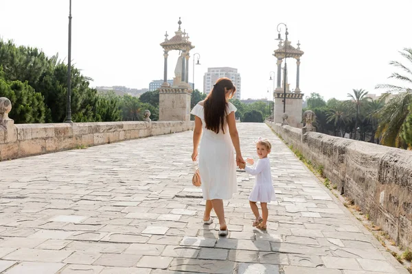 Женщина держит за руку ребенка в платье во время прогулки по мосту Пуэнте-дель-Мар в Валенсии — стоковое фото