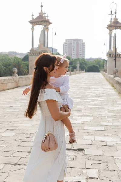 Vue latérale de la mère souriante en robe d'été tenant enfant sur le pont Puente Del Mar à Valence — Photo de stock