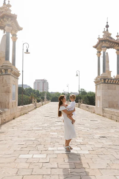Vista laterale della donna in abito bianco che abbraccia la figlia del bambino sul ponte Puente Del Mar a Valencia — Foto stock