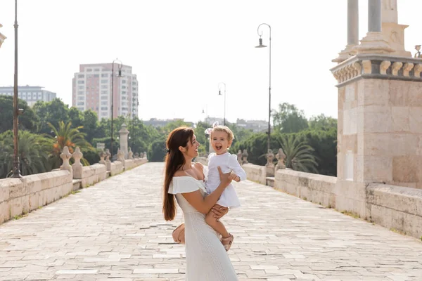 Mãe feliz no vestido de verão segurando bebê animado na ponte Puente Del Mar em Valência — Fotografia de Stock