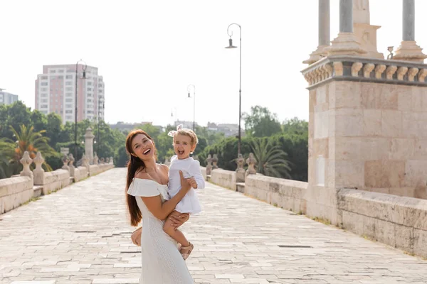 Happy woman holding baby in white dress and looking at camera on Puente Del Mar bridge in Valencia — Stock Photo