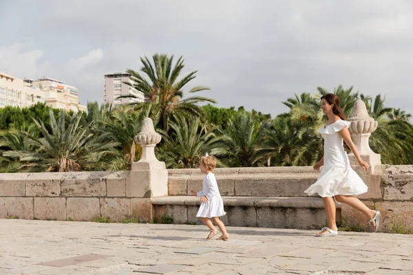 Vista lateral de mamá e hija en vestidos de verano corriendo en Puente Del Mar puente en Valencia - foto de stock