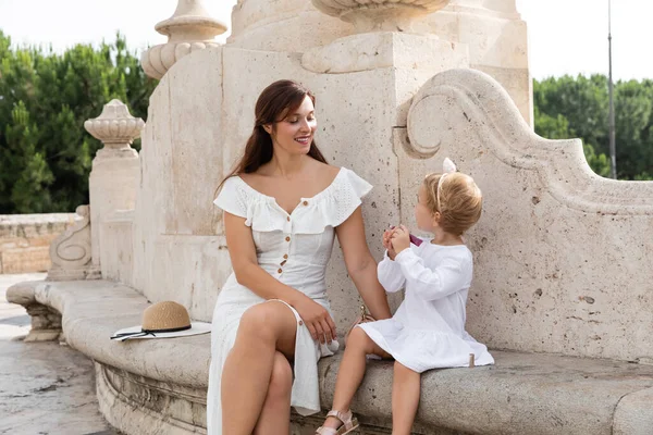 Baby girl holding lip gloss near mom and straw hat on bench of Puente Del Mar bridge in Valencia — Foto stock