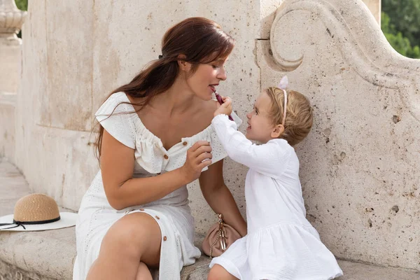 Side view of smiling child applying lip gloss on mother lips on stone bench of Puente Del Mar bridge in Valencia - foto de stock