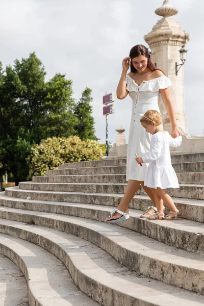 Cheerful mother walking near baby daughter with lip gloss on stairs of Puente Del Mar bridge in Valencia — Photo de stock