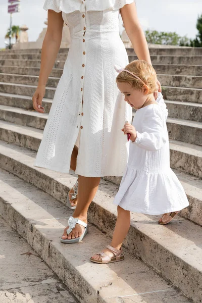 Baby girl holding lip gloss while walking near mom on stairs in Valencia - foto de stock