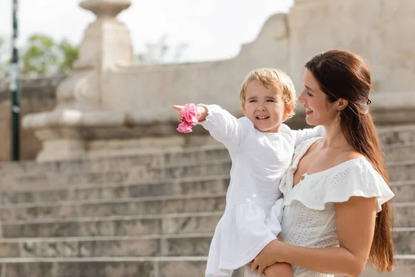 Cheerful baby girl holding flowers and pointing with finger near mom in summer dress in Valencia — Stockfoto