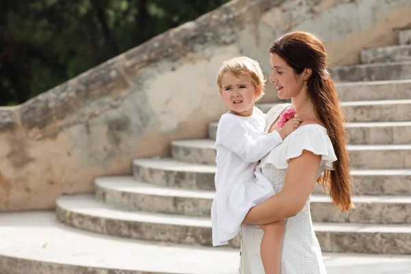 Fröhliche Frau im Sommerkleid hält verärgertes Baby mit Blumen in Valencia — Stockfoto