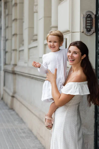 Smiling brunette woman in summer dress holding toddler girl near building in Valencia — Stock Photo
