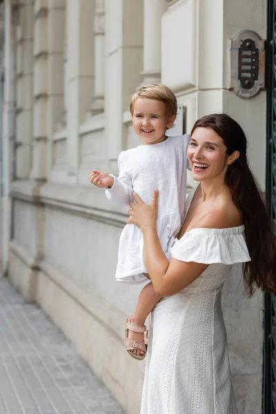 Mujer morena sonriente en vestido de verano sosteniendo niña pequeña cerca del edificio en Valencia - foto de stock