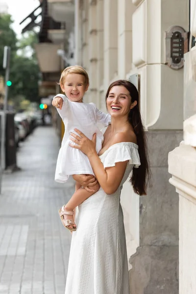 Cheerful brunette woman holding daughter pointing with finger on urban street in Valencia — Stock Photo