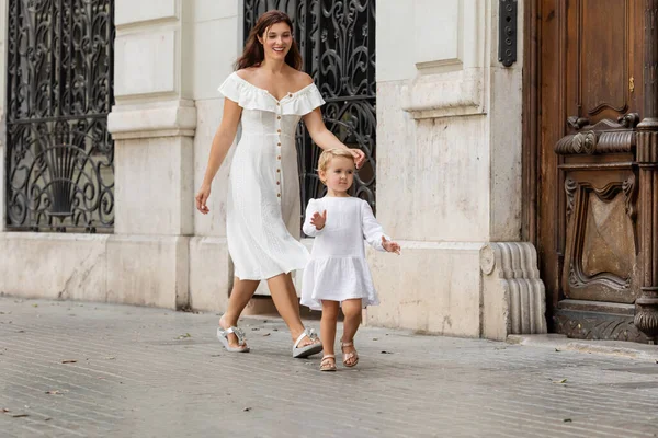 Niño pequeño caminando cerca de mamá sonriente en vestido de verano en la calle urbana de Valencia - foto de stock