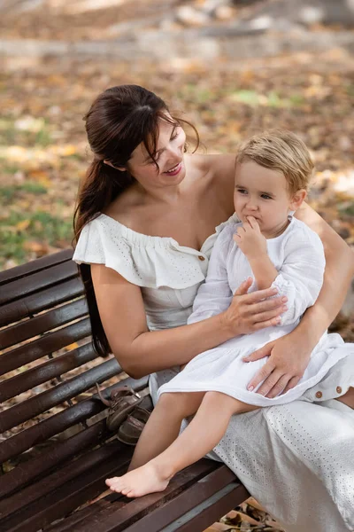 Smiling woman in dress hugging toddler daughter on bench in park — Stockfoto