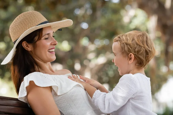 Маленькая девочка трогает улыбающуюся маму в шляпе в парке — стоковое фото