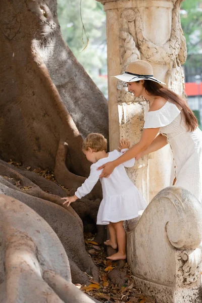 Smiling mother in sun hat touching baby daughter near wood outdoors in Valencia — Stock Photo