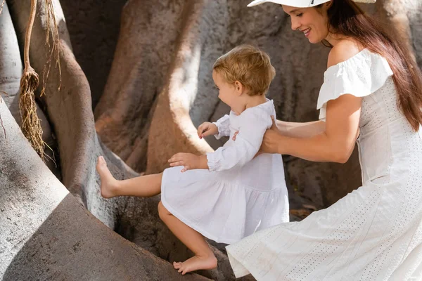 Side view of woman in dress holding daughter near tree outdoors in Valencia - foto de stock