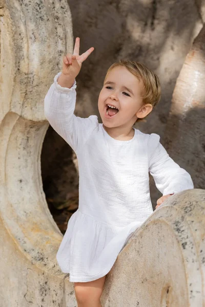Возбужденная девочка в платье показывает знак мира на каменной скамейке в Валенсии — стоковое фото