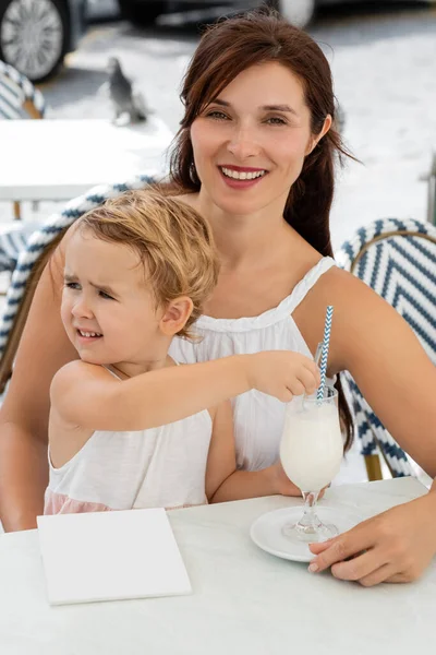 Улыбающаяся мать смотрит в камеру рядом с ребенком с молочным коктейлем в открытом кафе в Испании — стоковое фото