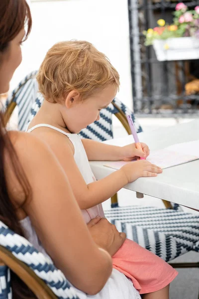 Vista lateral del dibujo del niño pequeño en el cuaderno cerca de la mamá en la cafetería al aire libre - foto de stock