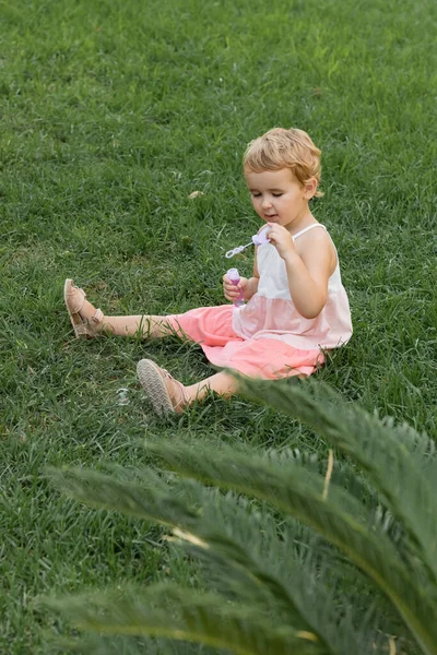 Малыш Тоддлер держит мыльные пузыри и сидит на траве на улице. — стоковое фото